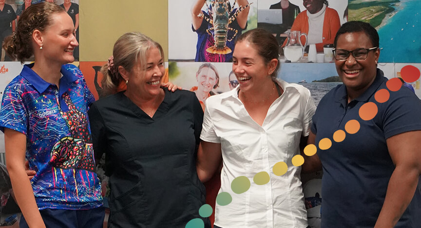 4 women, standing together arm in arm smiling, looking at each other.