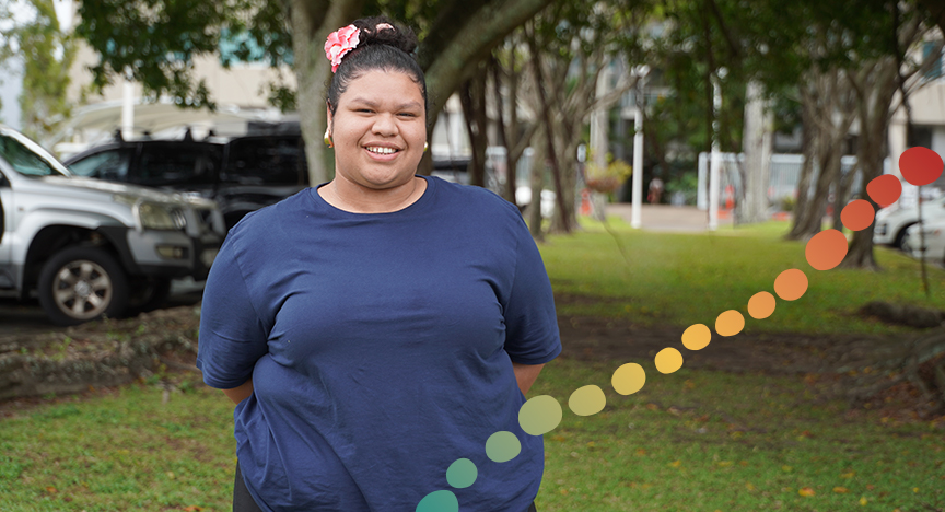 Smiling female standing on grass in front trees and parked cars.